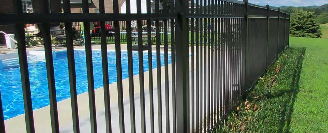 A black metal fence encloses a swimming pool next to a brick house under a blue sky. The scene highlights professional craftsmanship and durable materials, ideal for outdoor living and fence installation projects in Chesapeake.| First In Decks and Fencing | Virginia Beach Deck Builders