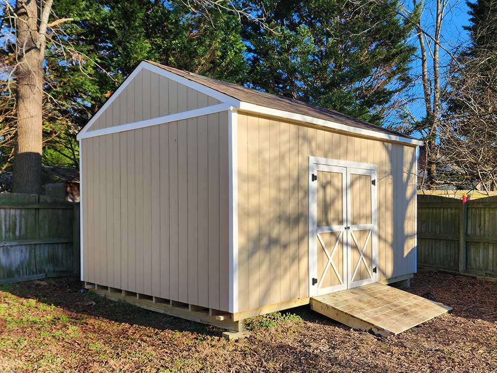 A beige shed with white trim and a wooden ramp sits in a fenced backyard, showcasing professional craftsmanship in outdoor structures as part of a backyard transformation project in Chesapeake.| First In Decks and Fencing | Virginia Beach Deck Builders