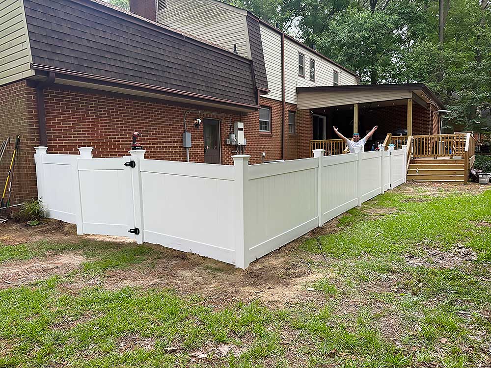 A person stands with arms raised next to a new white vinyl fence and a wooden deck attached to a brick house, showcasing professional craftsmanship in fence installation and outdoor living enhancements.| First In Decks and Fencing | Virginia Beach Deck Builders
