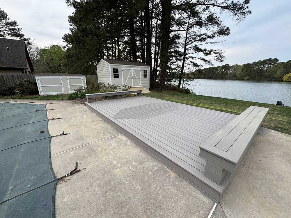 A spacious, light gray composite deck with a built-in bench overlooks a serene lakeside, framed by lush trees and a neatly painted shed, exemplifying high-quality deck installation for outdoor living in Norfolk.| First In Decks and Fencing | Virginia Beach Deck Builders