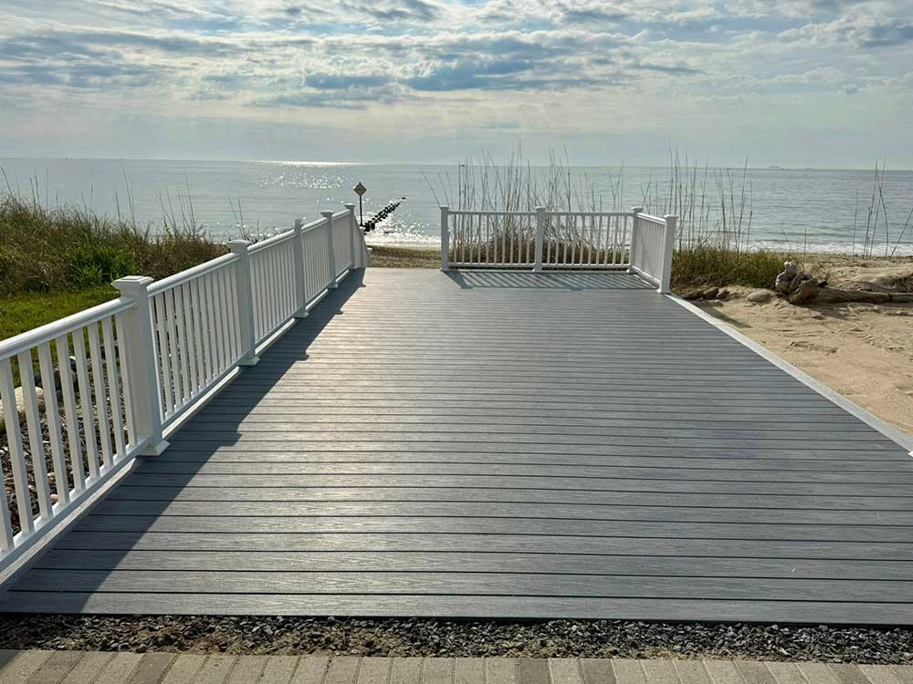 A spacious deck made of high-quality composite decking overlooks a serene beach, bordered by white railings. This outdoor living space showcases professional craftsmanship and durable materials, ideal for a backyard transformation.| First In Decks and Fencing | Virginia Beach Deck Builders