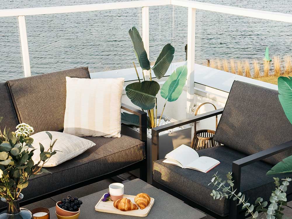 A cozy outdoor seating area with gray cushioned chairs, a white pillow, and a table set with croissants and coffee overlooks a body of water.…| First In Decks and Fencing | Virginia Beach Deck Builders