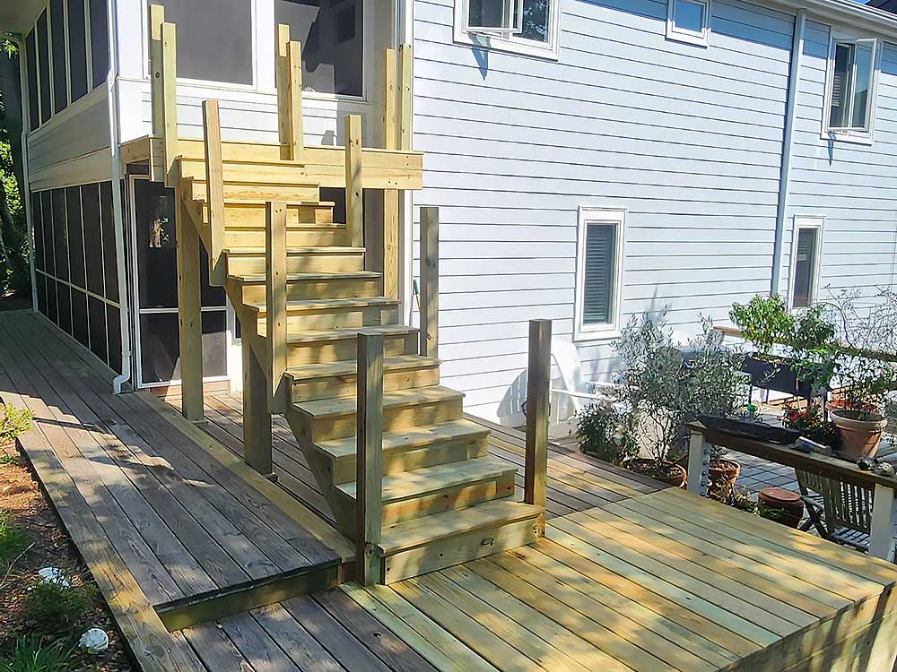 A newly installed custom deck with pressure treated lumber, featuring stairs leading to a second-story entrance. The light wood contrasts with the white siding of the house, enhancing the outdoor living space with high-quality craftsmanship.| First In Decks and Fencing | Virginia Beach Deck Builders