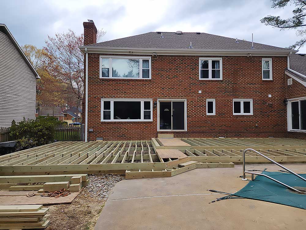 wood-deck-carousel-11 A new custom deck installation in progress in a backyard with pressure treated lumber framing, set against a two-story brick house, showcasing the foundation of an outdoor living space transformation in Chesapeake.| First In Decks and Fencing | Virginia Beach Deck Builders