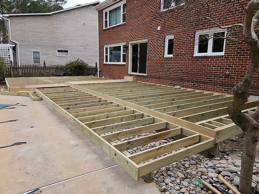 A partially constructed wooden deck framework is attached to a brick house, showcasing professional craftsmanship in deck installation with durable materials, ideal for enhancing outdoor living spaces.| First In Decks and Fencing | Virginia Beach Deck Builders