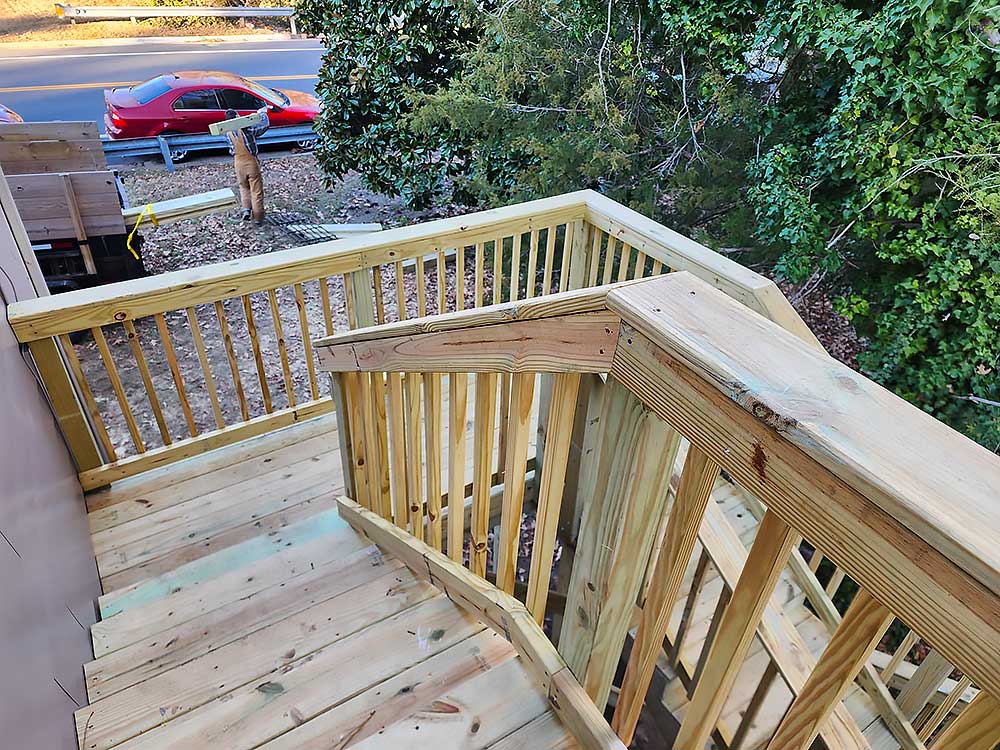 Wooden staircase leading to a backyard, surrounded by trees, showcasing high-quality, pressure treated lumber used in custom deck installation, with a worker carrying materials in the background, representing outdoor living craftsmanship.| First In Decks and Fencing | Virginia Beach Deck Builders