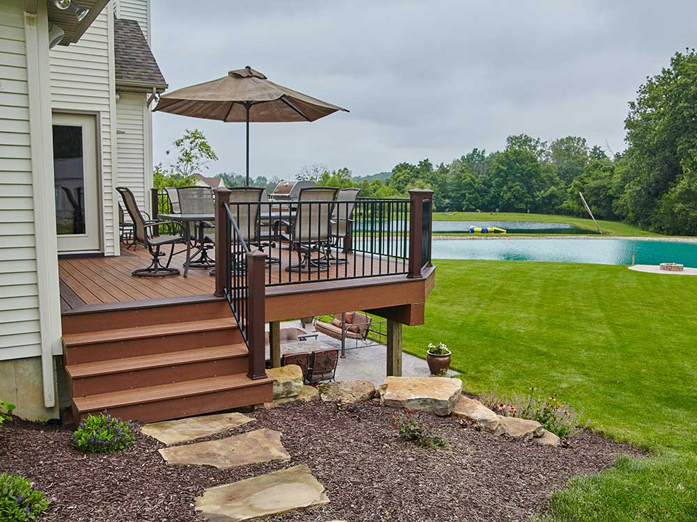 A spacious custom deck made of high-quality composite decking overlooks a lush backyard with a pond, featuring outdoor furniture and an umbrella, showcasing professional craftsmanship and promoting outdoor living.| First In Decks and Fencing | Virginia Beach Deck Builders