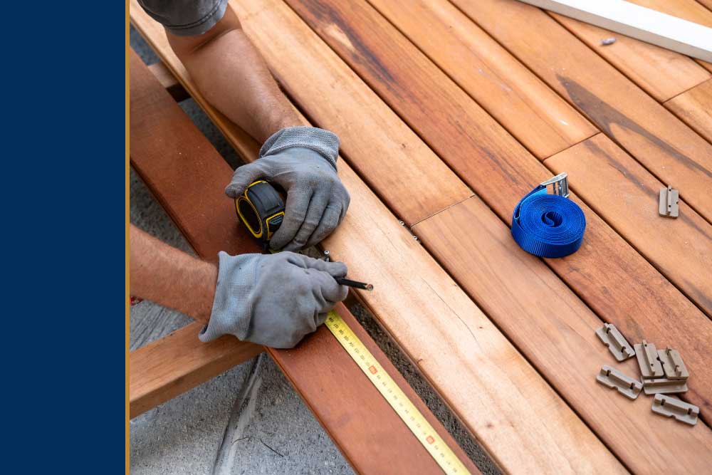 A person wearing gloves uses a tape measure and pencil to mark a wooden board during deck installation, emphasizing high-quality craftsmanship and durable materials for custom decks.| First In Decks and Fencing | Virginia Beach Deck Builders