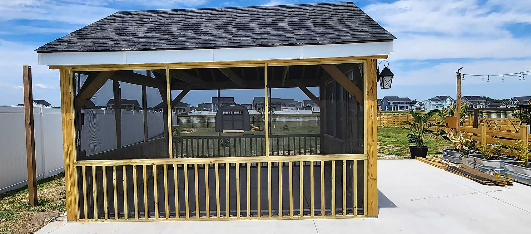 A wooden gazebo stands on a concrete patio, featuring a black shingled roof and screened sides, surrounded by plants and a wooden fence, illustrating professional craftsmanship and outdoor living in Chesapeake.| First In Decks and Fencing | Virginia Beach Deck Builders