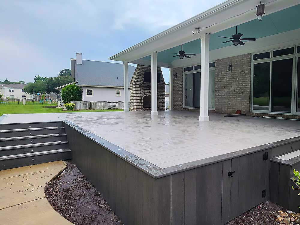 A spacious patio with a light gray composite decking and covered roof, featuring ceiling fans, adjacent to a brick fireplace. This outdoor structure exemplifies high-quality deck installation for enhancing outdoor living spaces.| First In Decks and Fencing | Virginia Beach Deck Builders