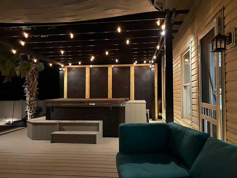 A cozy nighttime scene featuring a pergola illuminated by string lights, with a hot tub and comfortable seating on a deck made of durable materials, ideal for outdoor living and backyard transformation in Chesapeake.| First In Decks and Fencing | Virginia Beach Deck Builders