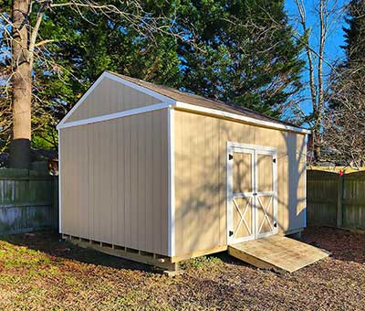 A beige shed with white trim stands in a backyard, surrounded by trees and a wooden fence. The shed features a double door and ramp, exemplifying high-quality outdoor structures suitable for storage, emphasizing backyard transformation.| First In Decks and Fencing | Virginia Beach Deck Builders