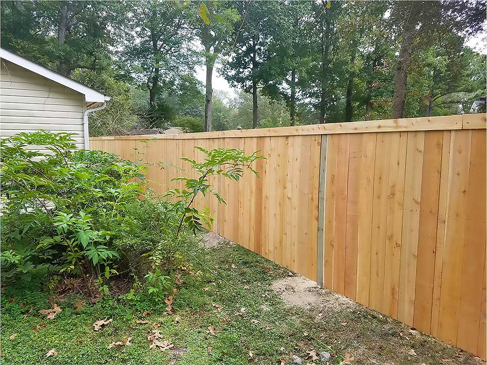 A newly installed wooden fence made of pressure treated lumber runs alongside a house, surrounded by green foliage, demonstrating professional craftsmanship and enhancing outdoor living space in a backyard transformation.| First In Decks and Fencing | Virginia Beach Deck Builders