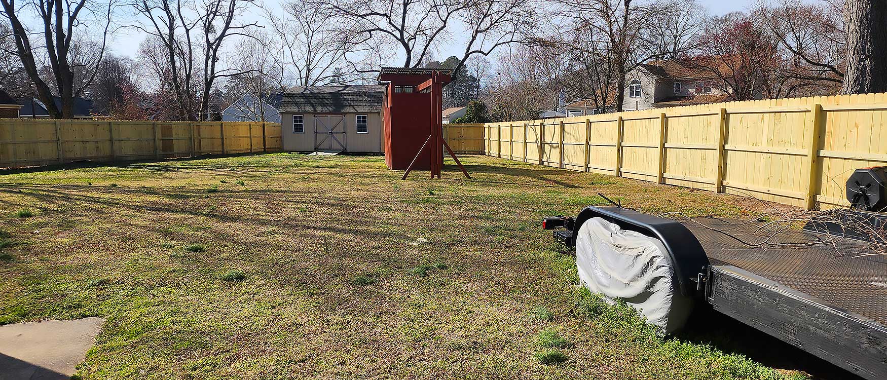 wood-fences-page-long A spacious backyard features a well-maintained lawn, a wooden play structure, a large shed, and a new wooden fence installation, emphasizing outdoor living and home improvement with durable materials.| First In Decks and Fencing | Virginia Beach Deck Builders